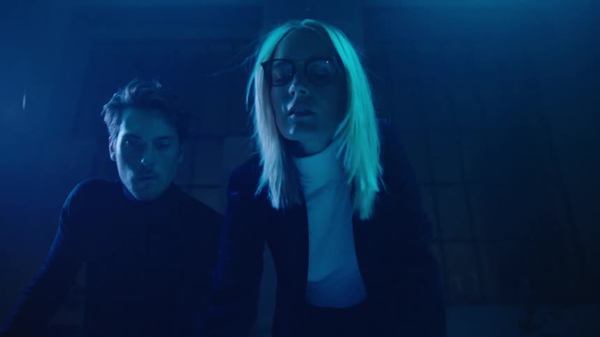 Woman with blonde hair wearing glasses and holding a marker leans forward as a man stands next to her