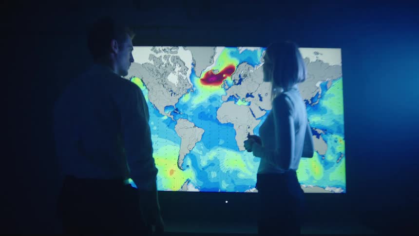 Man points to a weather map on a television as a blonde woman stands next to him