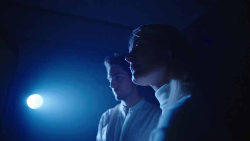 Woman with fair hair white turtleneck sweater stands next to man wearing white shirt in spotlight looking at big screen computer in dim lit data center