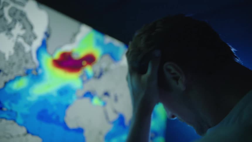 A man is standing in front of a large television looking at weather radar and he rubs his forehead