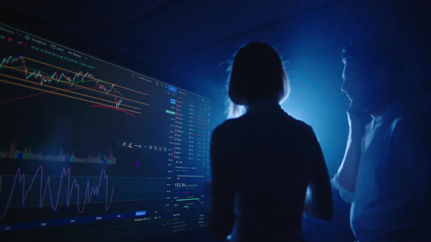 Man and woman stand in dark room lit by single bright light discussing graphs on large digital display