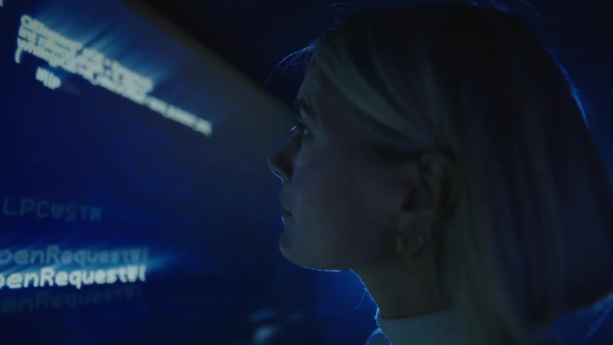 Man and woman stand examining computer code displayed on large digital screen in shadowed room lit by bright blue light