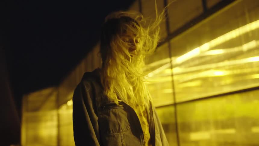 Blonde woman with denim jacket stands in wind next to building with flickering yellow lights at night
