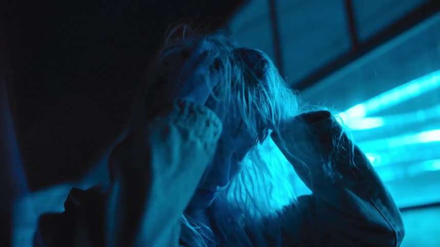 A woman looking down with both hands in her hair rubbing and tugging while colorful lights flash and strobe