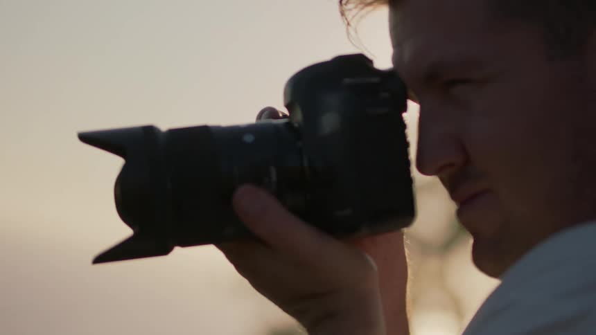 Man stands outdoors near water focusing through camera on sunny day