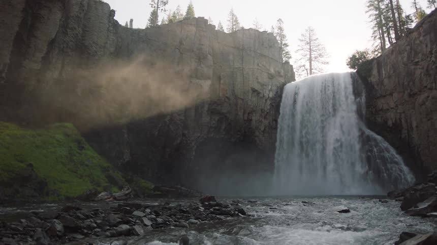 Waterfalls flow into a creek as steam rises and there are rock formations with trees in the distance