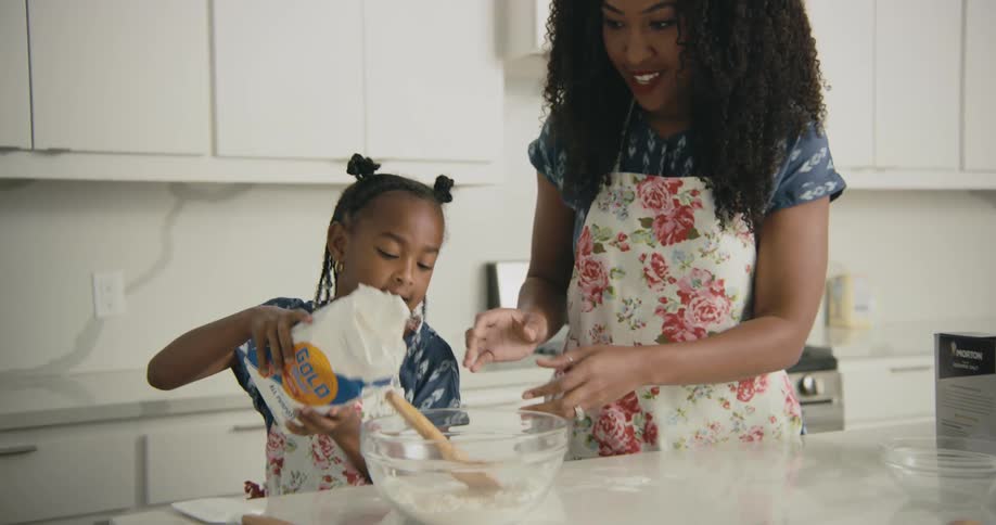 Mother opens a bag of flour and her daughter pours the flour into a glass bowl and spills some on the counter