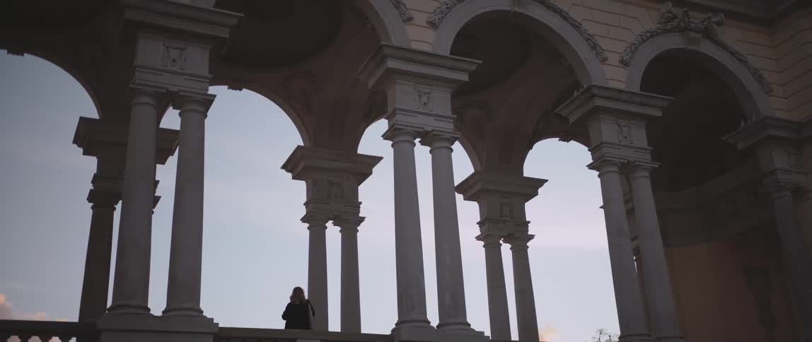 Woman with a purse over her shoulder stands next to a pillar under an arch of a building under a blue sky