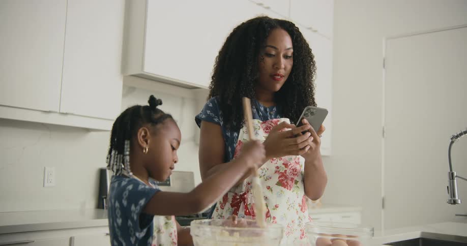 Mother looks at her cell phone then puts it in a pocket while her daughter mixes dough in a glass bowl with a spatula