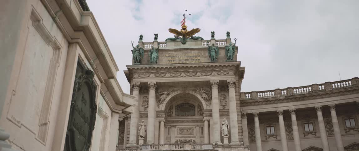 Birds and a flag fly over a large building with statues and ornate designs under a cloudy blue sky