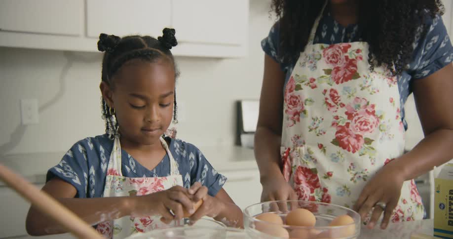 Young girl wearing an apron with flowers on it cracks a brown egg into a bowl as her mother stands next to her in the kitchen
