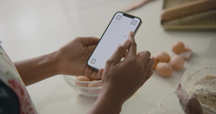 A woman scrolls through her smartphone while her daughter mixes flour in a bowl