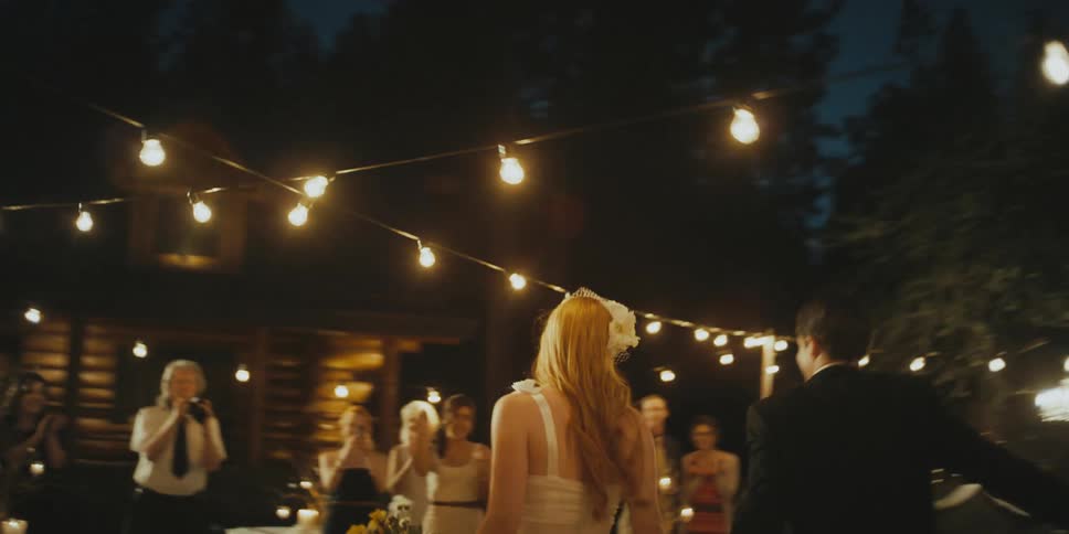 A bride and groom are walking to a crowd of people with strings of lights up