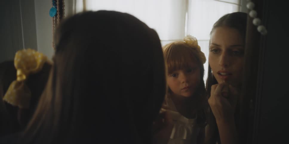 Little girl watches while woman puts lipstick on her lips in mirror