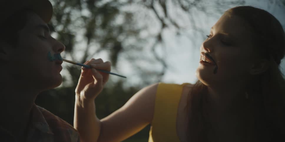 A woman is painting a fake mustache on a man as they sit outdoors