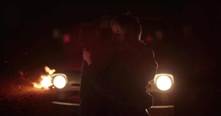 Couple embracing in front of a vehicle with its headlights on next to a campfire
