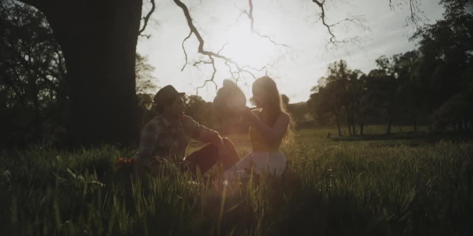 A man and woman are sitting in the grass beneath a tree playing with their hats