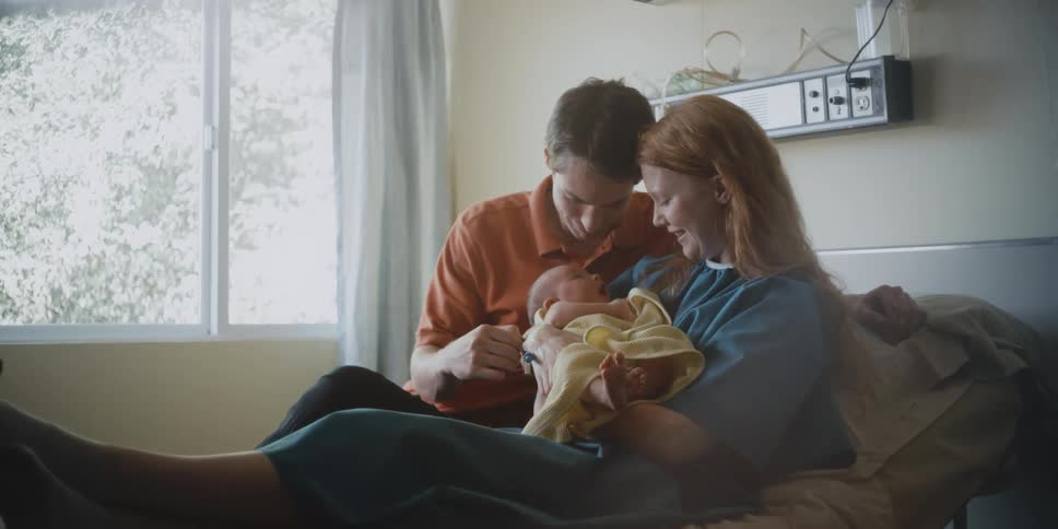 Man and woman sit in bed holding an infant