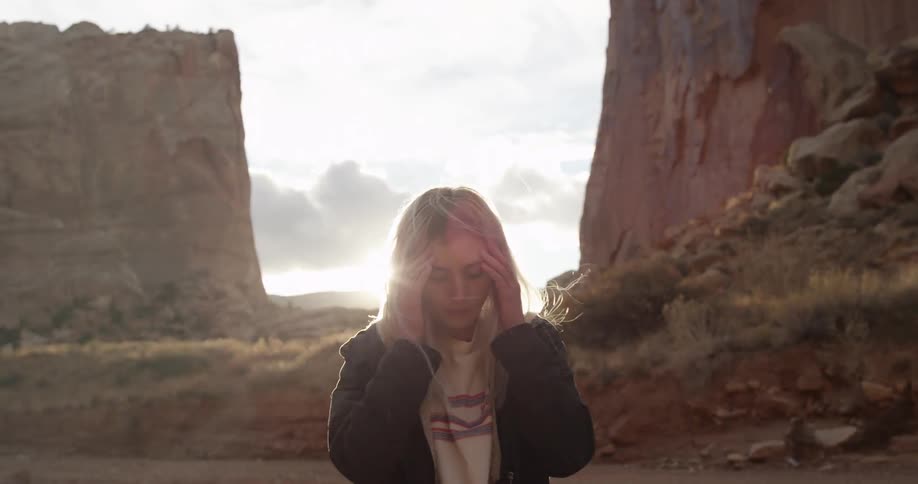 Blonde woman brushing back her hair while standing between two large rocks at sunset