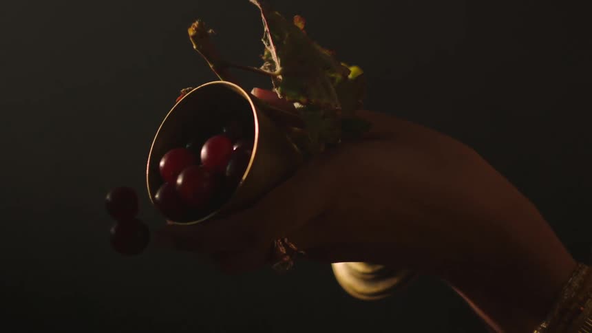 A womans hand can be seen pouring whole grapes from a golden goblet