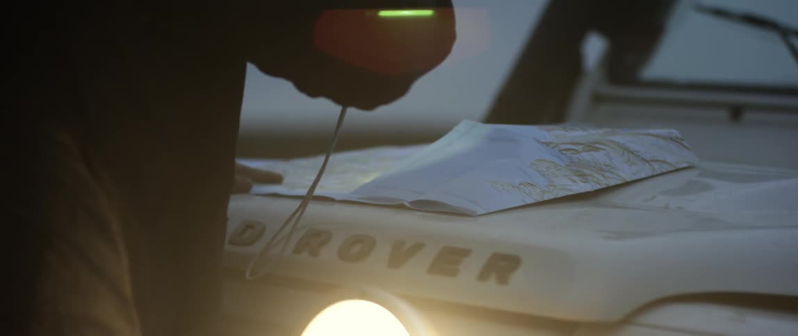 A man holds a flashlight trying to hold down and read a map on the hood of his land rover