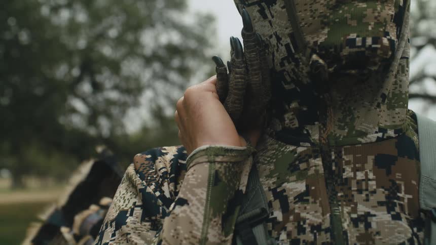 Person wearing camouflage clothing holds black fingers with claws on his shoulder while near the green trees