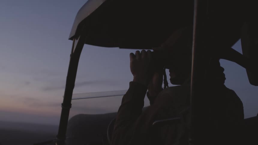 A person looks out at the horizon from a vehicle using a pair of binoculars at sunset