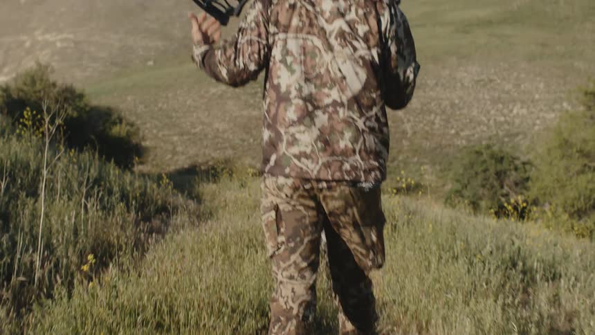 Bow hunter wearing camouflage walks through grass toward large hill carrying bow and arrows around against the back of his neck
