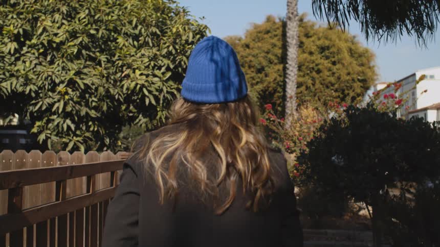 Woman with blue wooly hat brown wavy hair and black overcoat walks down the street