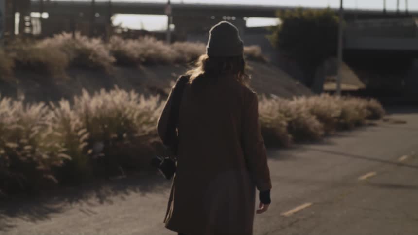 Woman with gray wooly hat brown hair and coat walks down road carrying camera