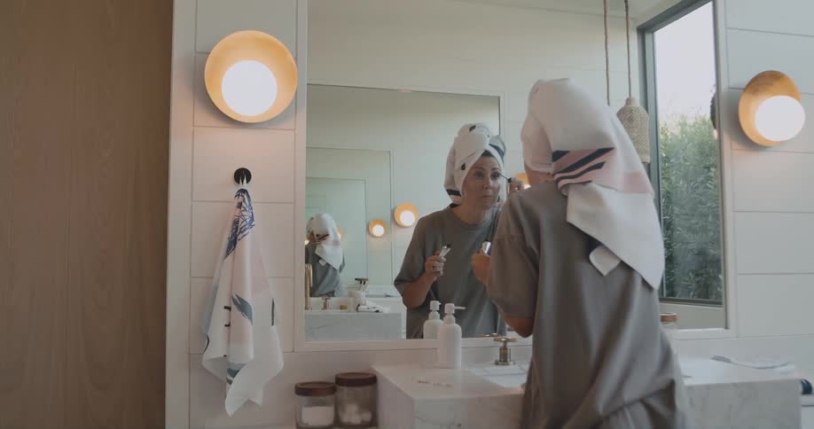 Woman stands in bathroom putting on mascara while looking into the mirror