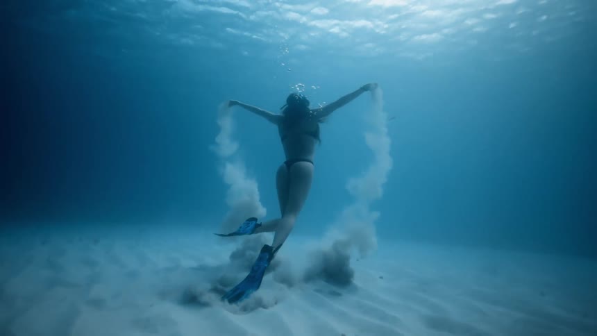 A woman picks up sand from the ocean bottom and lets it go as she swims up toward the surface