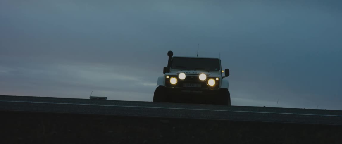 Under a very cloudy sky a large white jeep drives up an embankment and onto a paved road