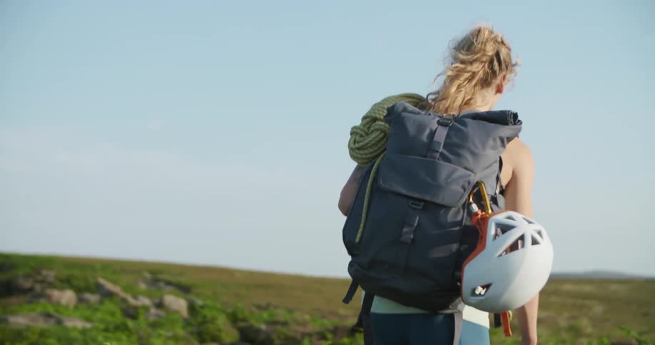 A woman wearing a backpack and carrying climbing gear hikes through a field