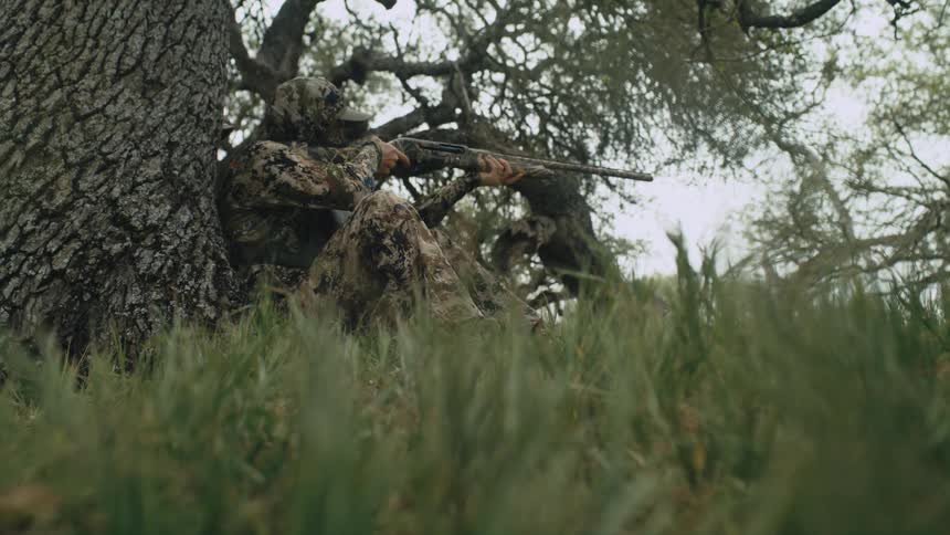 A hunter is pointing his weapon while sitting by a tree