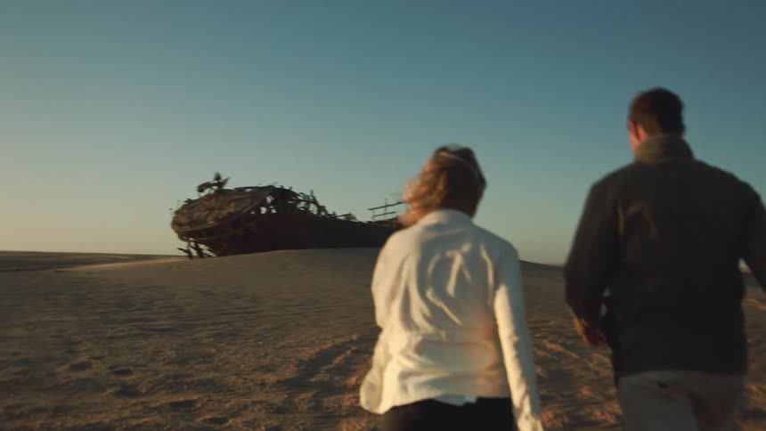 Man and woman walk toward desert shipwreck with excitement