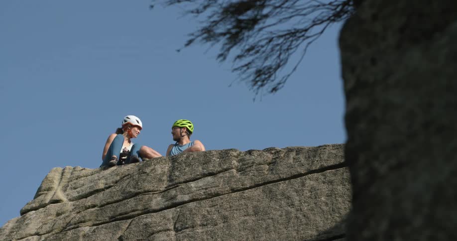 Two rock climbers sit near edge of cliff wearing helmet under a clear blue sky