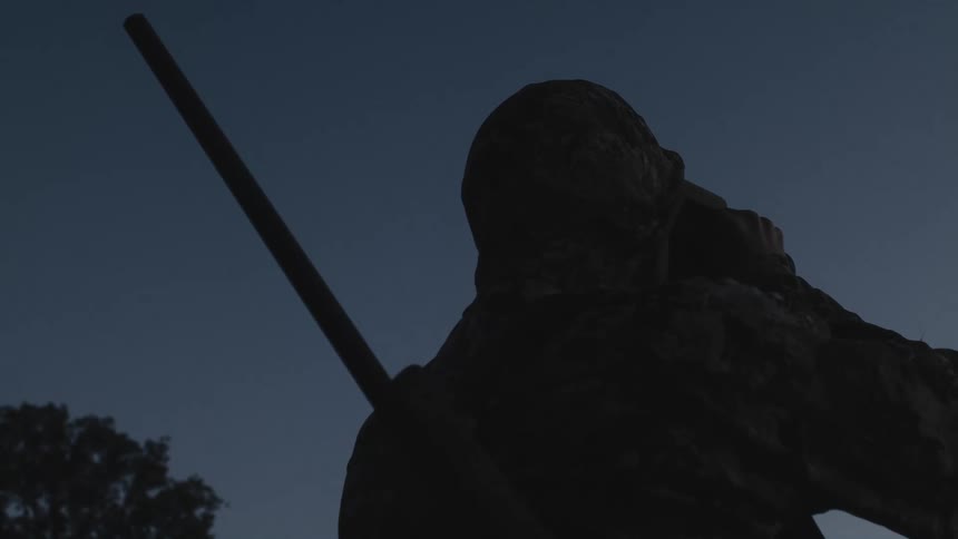 Man with shotgun looks at horizon through binoculars at dusk