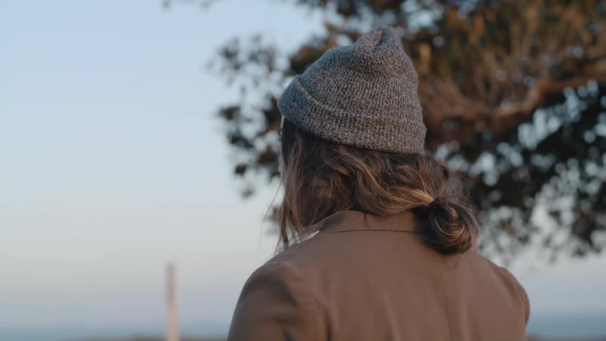 Woman with gray wooly hat brown wavy hair and jacket walks next to tree in the field