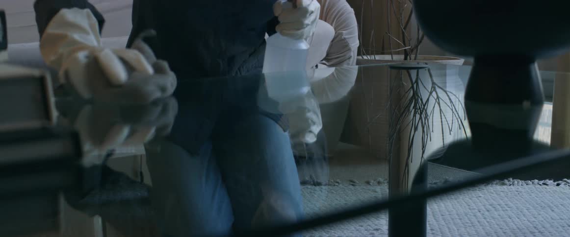 A woman wearing gloves and a mask polishes a glass coffee table