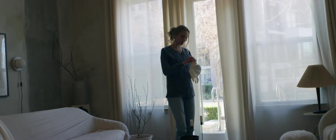 A woman stands in her living room and puts on gloves for cleaning while she lightly shakes her hips and head to a rhythm.