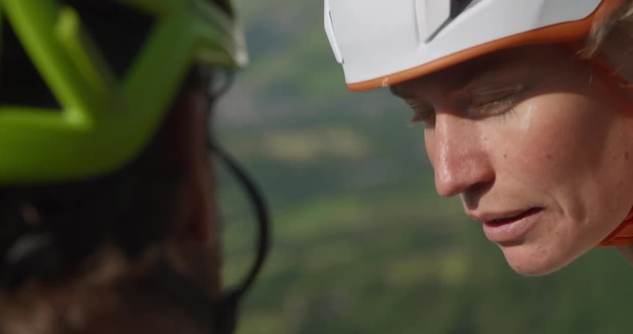 Two women chat while standing on a hill wearing helmets overlooking a field