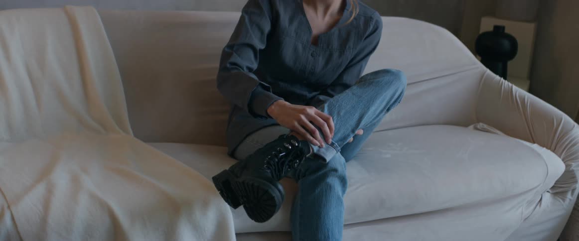 A woman sits on a couch at home and rubs her lower leg and then stretches her arms out in front of her as she releases a deep breath of air