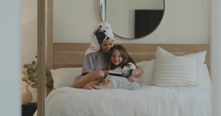 Mother with hair towel and gray shirt reads book with daughter lying on bed in bedroom