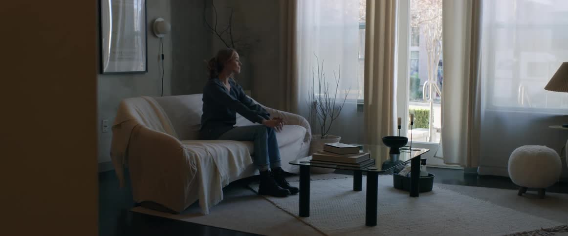 A woman sitting on a couch stands up and walks over to an item that she picks up from the floor