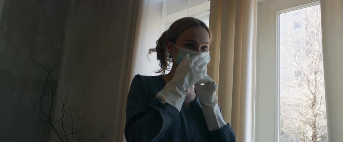 Woman with blue shirt white gloves and jeans puts on mask in living room at home