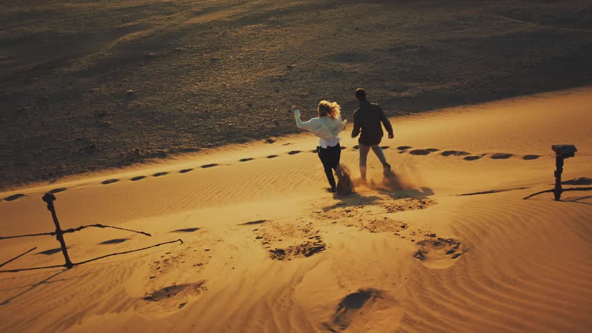 Man and woman playfully run down a sand dune at sunset