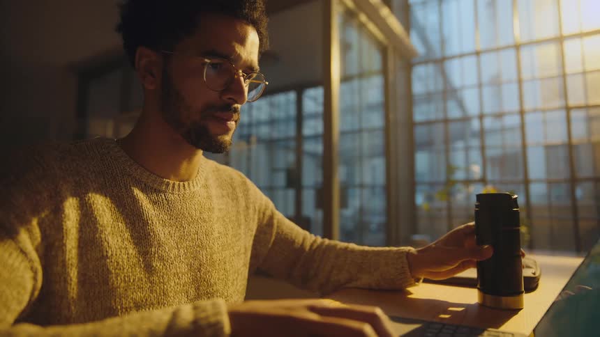 A man drinks from a thermal mug while using a laptop at a desk in the evening