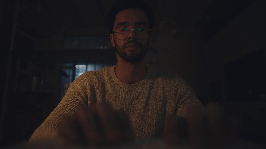 Tired man with glasses beard and gray sweater uses laptop in dark room