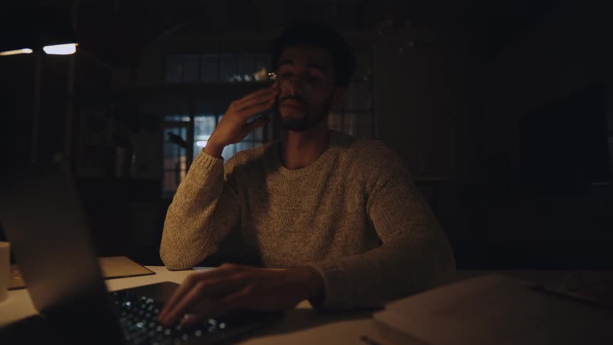 Man typing on laptop in dark room rubs his eyes and forehead